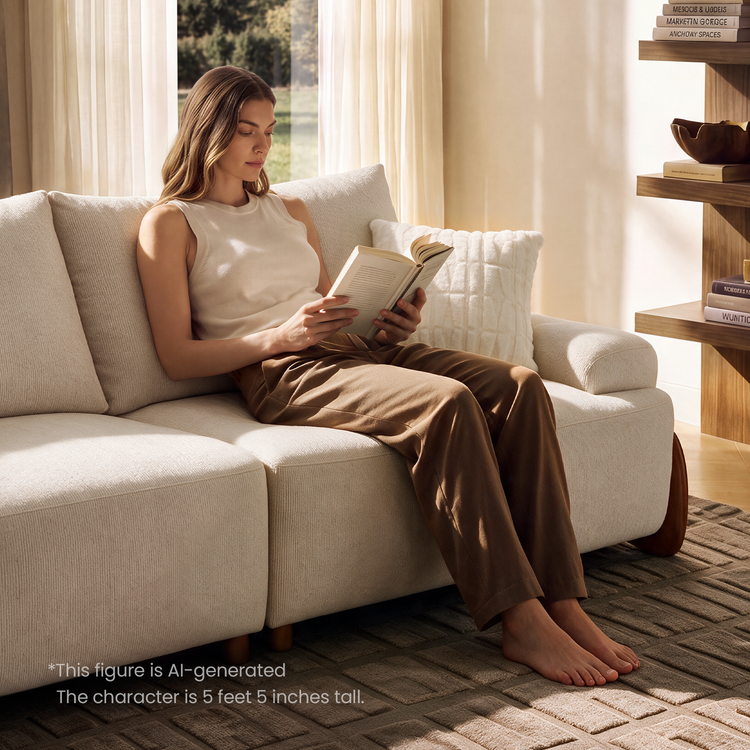 A woman sits barefoot on a cream sofa reading an open book, with a white pillow beside her and sunlight coming through curtains.