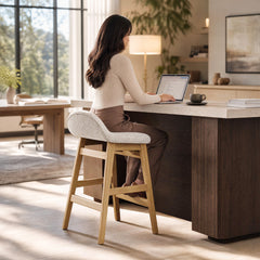 A woman uses a laptop at a counter while seated on a tall, light-colored stool with a curved upholstered back, angled legs and footrests.