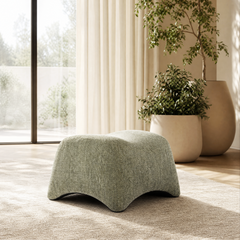 Low green upholstered ottoman has a sculpted wavy profile, Ottoman sits on a rug near large windows