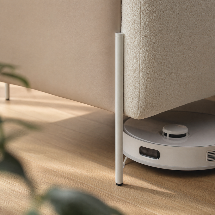 A white robot vacuum sits beneath cream upholstered furniture beside a slender white leg on a light wood floor.