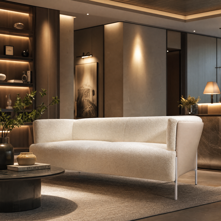 Cream-colored textured sofa with a curved back, raised sides and slender legs in a warmly lit room with a round coffee table, decorative vases and illuminated shelving.