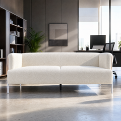 Cream textured sofa with two seat cushions, rounded arms and slender legs in a sunlit office with shelving, plants and a desk behind it.