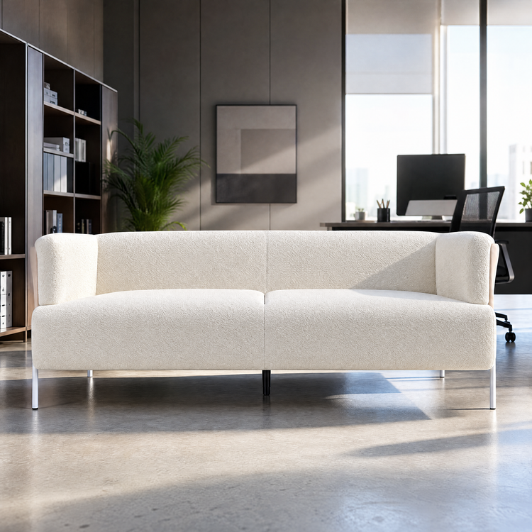 Cream textured sofa with two seat cushions, rounded arms and slender legs in a sunlit office with shelving, plants and a desk behind it.
