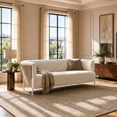 Cream sofa with a taupe cushion and fringed throw on a textured beige rug in a sunlit room with tall curtained windows, a side table, lamp and low brown cabinet.