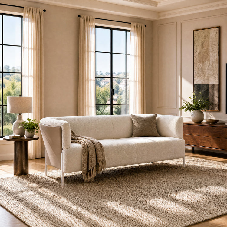 Cream sofa with a taupe cushion and fringed throw on a textured beige rug in a sunlit room with tall curtained windows, a side table, lamp and low brown cabinet.