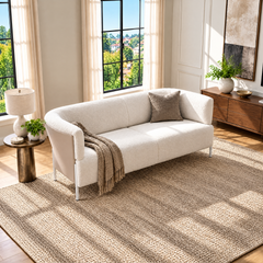 A curved white sofa with a taupe cushion and fringed throw sits on a textured beige rug in a sunlit room with tall windows, a side table, lamp and brown cabinet.