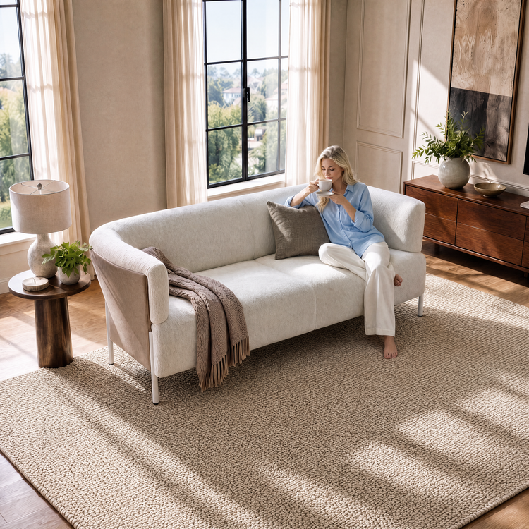 A woman holds a cup and saucer on a curved cream sofa with a brown throw and cushion in a sunlit room with tall windows, a textured beige rug, plants and a sideboard.