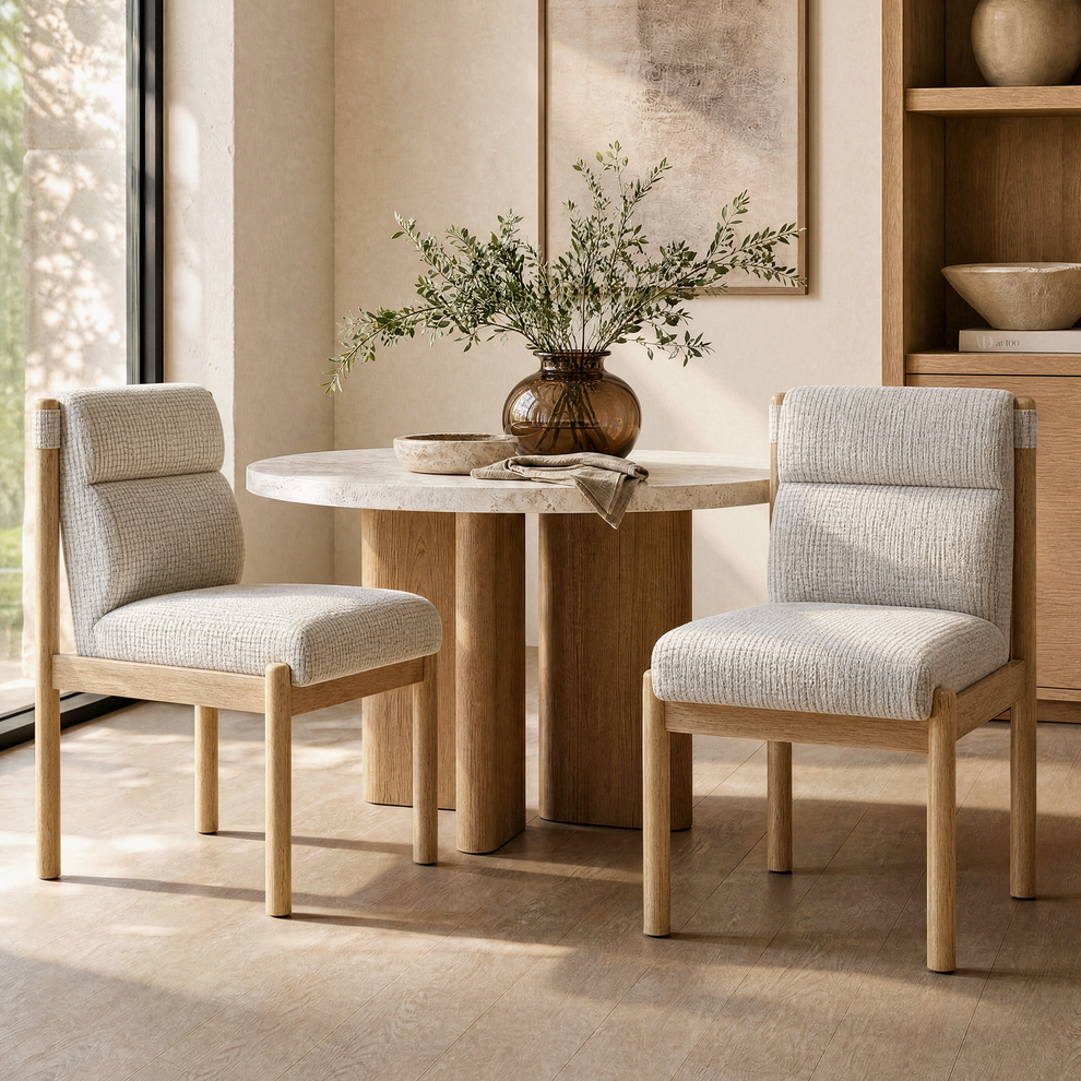 Two cream upholstered chairs with light brown frames flank a round pedestal table topped with a brown vase of leafy branches, a shallow bowl and a draped cloth.