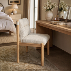 A cream upholstered chair with a light wood frame sits beside a wooden vanity in a bedroom; a vase of white flowers, perfume bottles, and jewelry are arranged on the vanity.