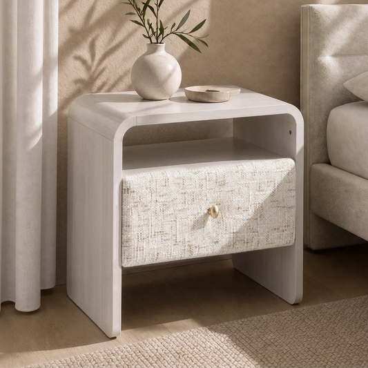 A light-colored bedside table with a woven drawer and open shelf stands beside a bed, topped with a vase of leafy branches and a shallow dish.