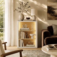 A white open bookcase glows with warm interior shelf lighting, Books and ornaments fill three tan-lined tiers