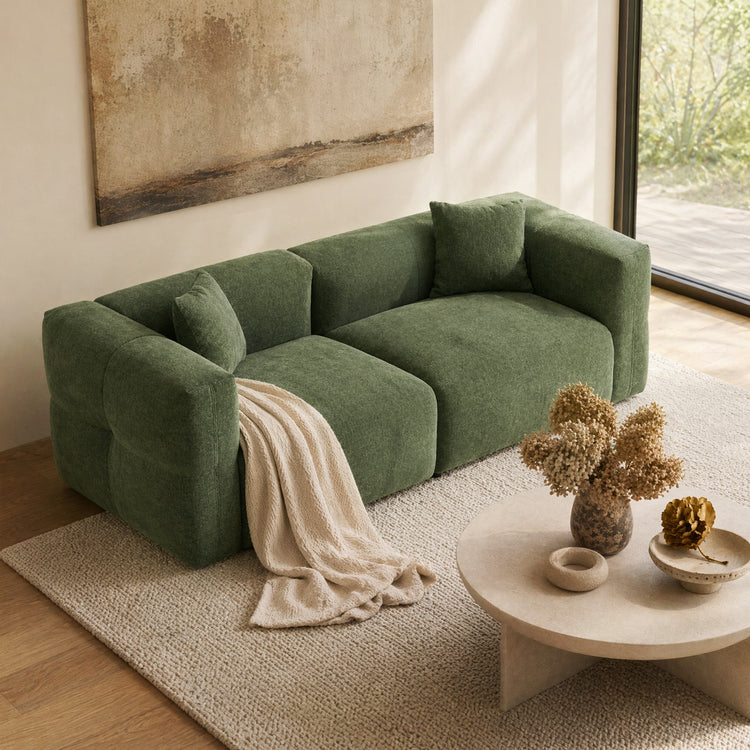 Green upholstered sofa with two cushions and a cream throw in a sunlit living room.
