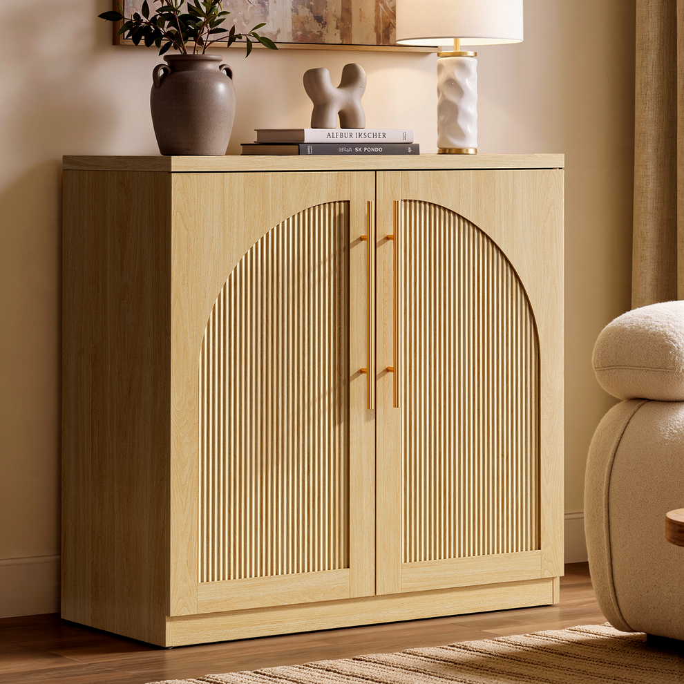 Light wood-tone cabinet with arched fluted double doors and gold-tone handles beside a cream upholstered chair