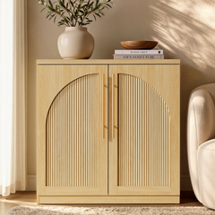 Light wood-grain accent cabinet with arched fluted double doors and long gold-tone handles beside a cream chair