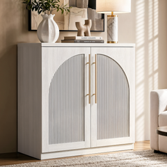 White accent cabinet with arched fluted double doors and long gold-tone handles beside a cream chair