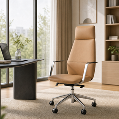 Tan high-back office chair with a padded headrest, silver-colored arm supports and wheeled base beside a dark desk in a bright room with large windows and shelving.