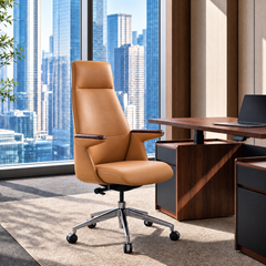 Tan high-back office chair with padded armrests and a silver five-spoke wheeled base beside a desk with a laptop, against tall windows overlooking city buildings.