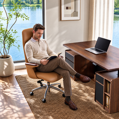A man reads a book in a tan high-back swivel chair beside a brown desk with an open laptop, near large windows overlooking water and trees.