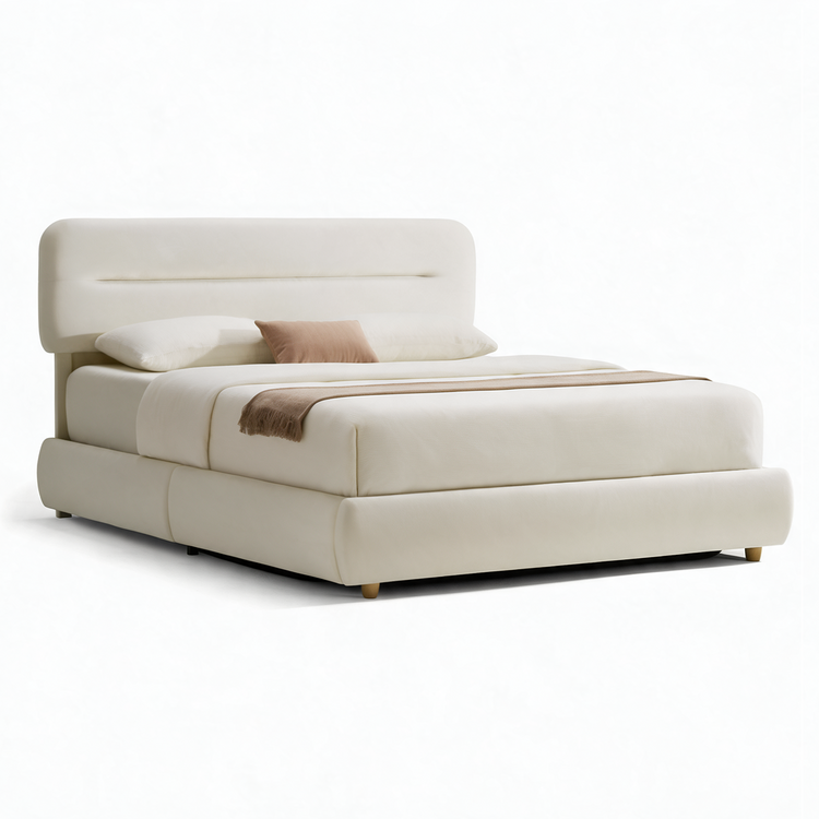 Cream-colored bed with a rounded padded headboard, matching low frame, white bedding, two white pillows, a tan accent cushion and a brown fringed throw across the foot.