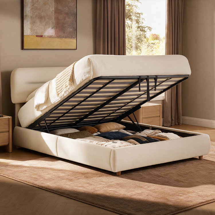 Cream upholstered bed with its mattress and slatted base lifted to reveal a storage compartment filled with pillows and blankets.