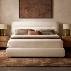 A cream upholstered bed with a rounded headboard is centered between wooden nightstands, with striped and rust-colored pillows, matching lamps, and a framed painting above.