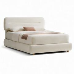 Cream-colored bed with a rounded rectangular headboard, padded frame, short legs, white bedding, a pink accent pillow and a fringed brown throw across the foot.