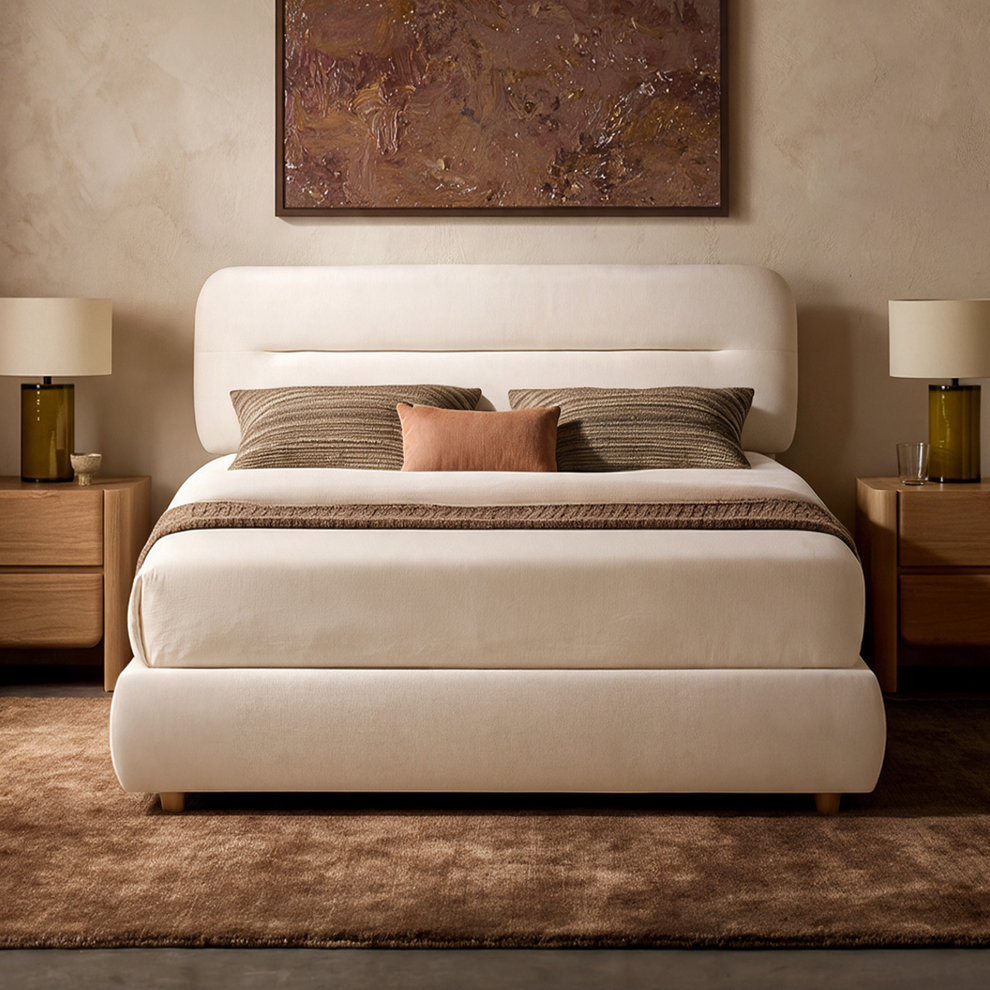 Cream upholstered bed with a rounded headboard, brown pillows and throw, flanked by bedside cabinets and lamps beneath a brown abstract painting.