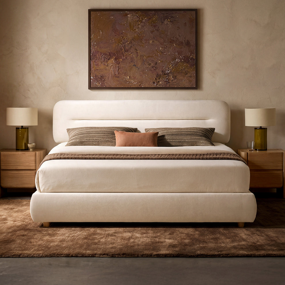 Cream upholstered bed with a rounded headboard, brown pillows and throw, flanked by bedside tables and lamps beneath a framed abstract painting.