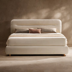 Cream upholstered bed with a rounded rectangular headboard, low curved base, pale bedding, two striped pillows and a smaller brown cushion against a beige wall.