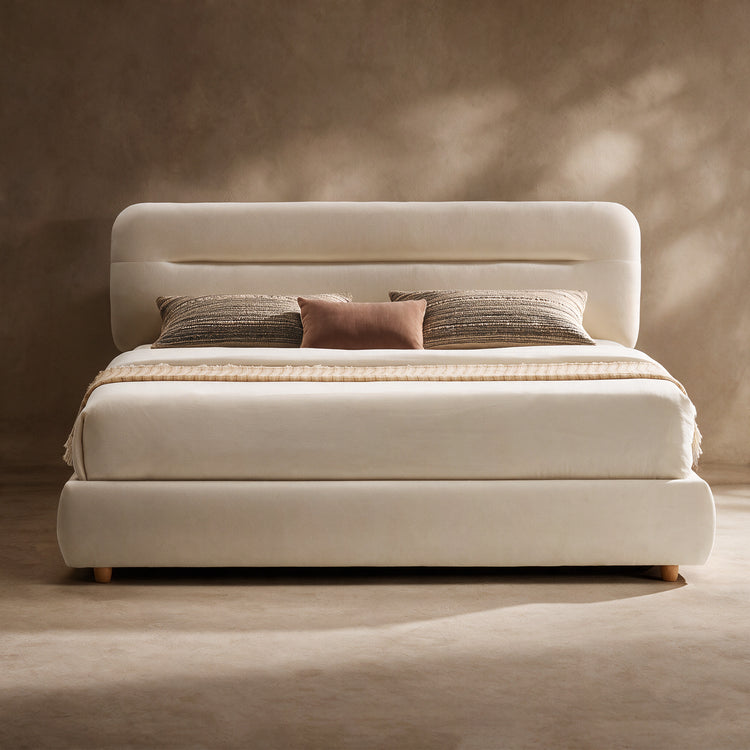 Cream upholstered bed with a rounded rectangular headboard, low curved base, pale bedding, two striped pillows and a smaller brown cushion against a beige wall.