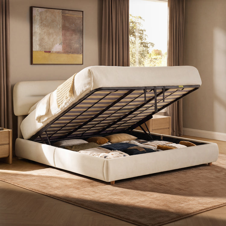 Cream upholstered bed with its mattress and slatted base lifted to reveal a storage compartment filled with cushions and folded bedding.