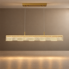 Illuminated gold-toned linear pendant light with rows of circular details, suspended by two thin cables from a rectangular ceiling mount above a beige surface.