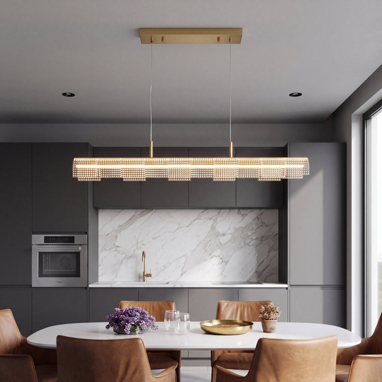 An illuminated gold-toned linear pendant with dotted rectangular panels hangs above a white dining table and tan chairs in a kitchen with dark gray cabinets.