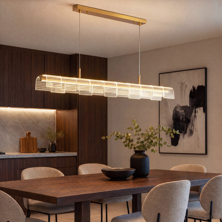 An illuminated linear pendant with gold-colored fittings and overlapping dotted panels hangs above a dark dining table surrounded by beige chairs.