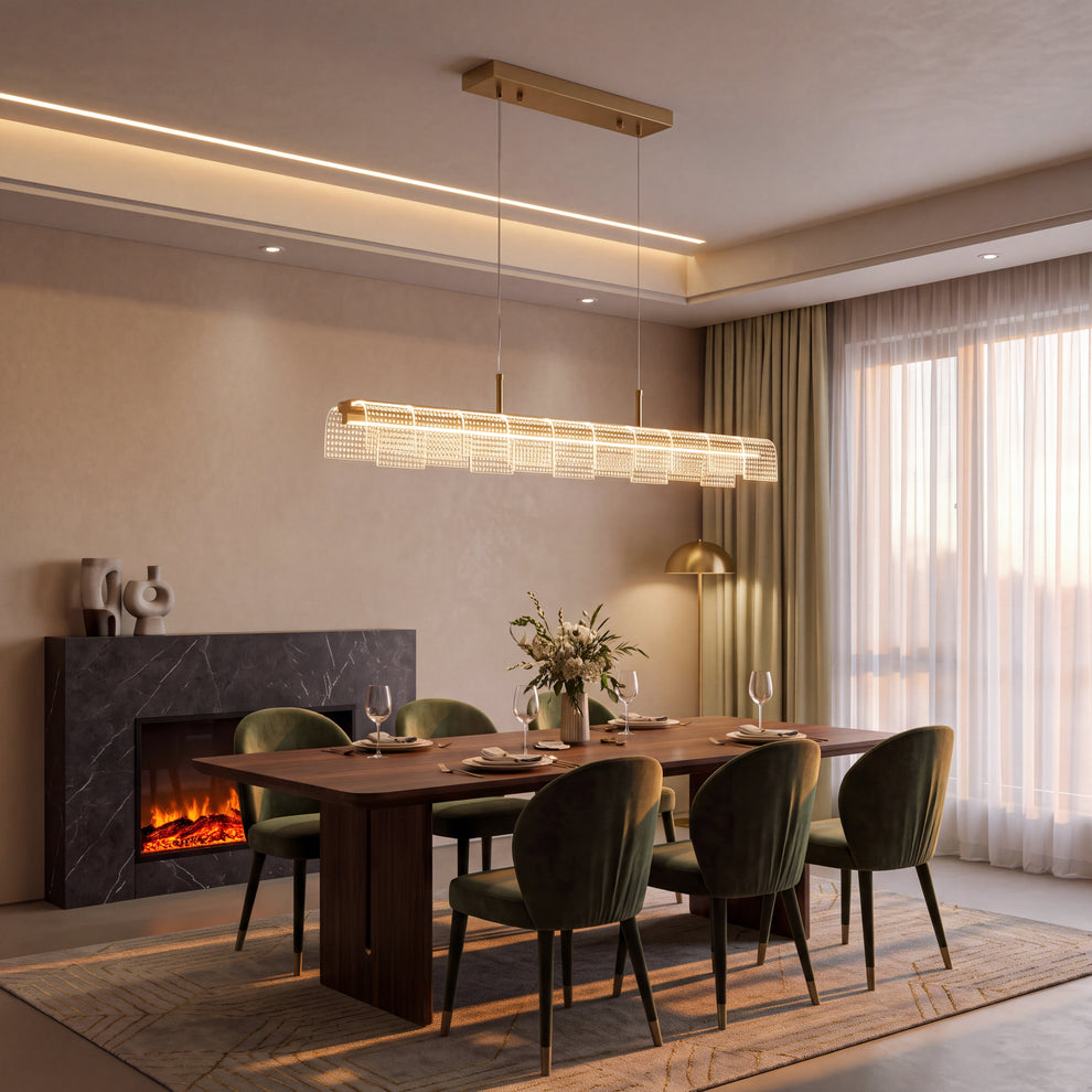 A long illuminated pendant with gold-toned trim and textured clear panels hangs above a set dining table with green chairs, beside a fireplace and tall curtained windows.