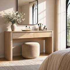 Light wood-grain table with two drawers stands beneath a rounded rectangular mirror, Flowers, cosmetics, and brushes decorate the sunlit tabletop