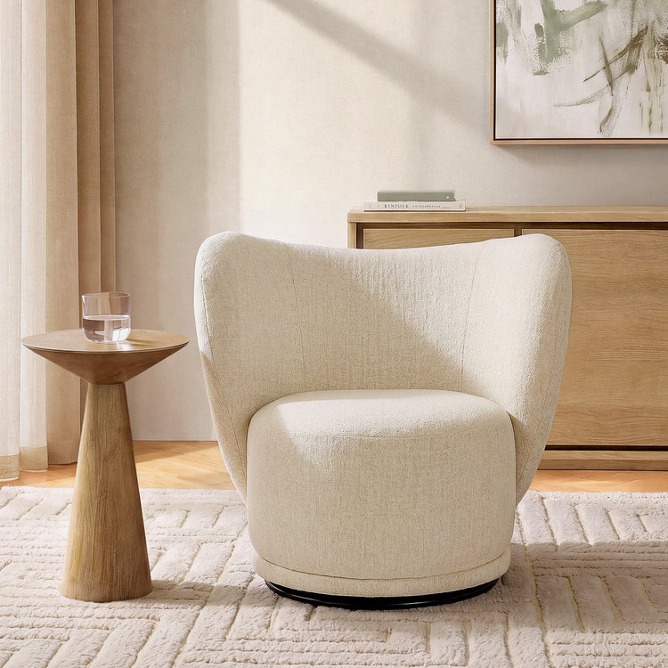 A cream wraparound chair with textured upholstery stands beside a wood pedestal table in a sunlit room