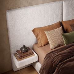 Close bedroom view of a tall cream upholstered headboard, brown bedding, pillows, and a small bedside shelf