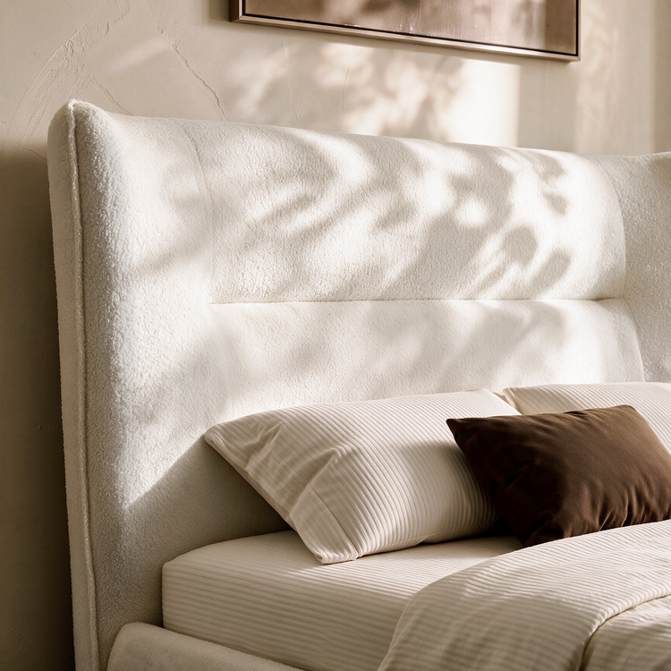 Close-up of a bed with a tall cream upholstered headboard, light striped bedding, and cream and dark brown pillows.