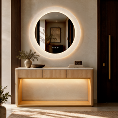 A round illuminated mirror hangs above a light wood console table, with a vase, bowl, and stacked books on top.
