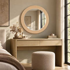 A round mirror with a textured frame hangs above a light wood vanity, with flowers, small containers, and a cushioned stool in a sunlit room.