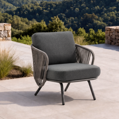 A gray woven outdoor lounge chair with a thick dark cushion stands on a sunny stone terrace