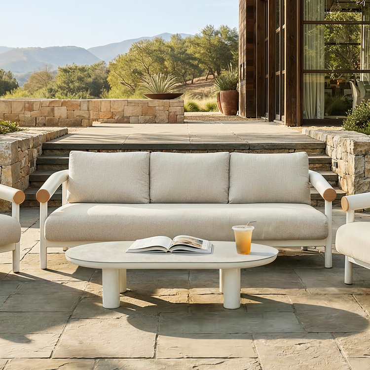 White cushioned outdoor sofa with wood arm accents beside a low oval table on a stone patio