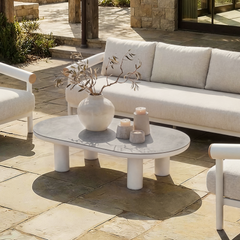 A light outdoor seating group surrounds an oval coffee table with a vase and candles on a sunlit patio