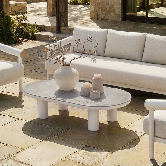 A light outdoor seating group surrounds an oval coffee table with a vase and candles on a sunlit patio