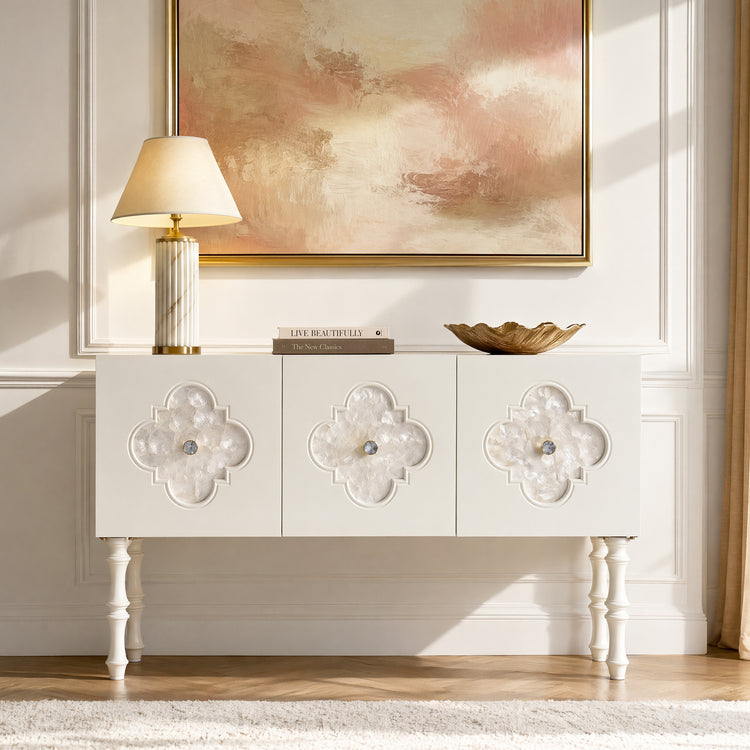 White three-door console with quatrefoil insets supports a lamp, books, and sculptural bowl beneath abstract artwork