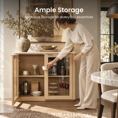 A woman reaches for a cup beside an open wooden cabinet stocked with coffee equipment, dishes, and glassware.