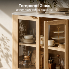Light wood-toned cabinet with transparent doors, one open, displaying stacked dishes, cups, a tiered stand and coffee accessories beneath a pale top with a vase and bowl.