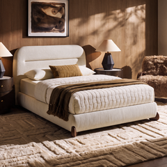 Cream upholstered bed with white quilted bedding and a brown throw, flanked by dark bedside tables and lamps in a sunlit bedroom with abstract wall art and a textured rug.