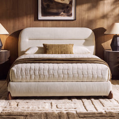 Cream upholstered bed with a rounded headboard, white quilted bedding, a brown throw and accent cushion, flanked by dark bedside tables and lamps against a wood-toned wall.